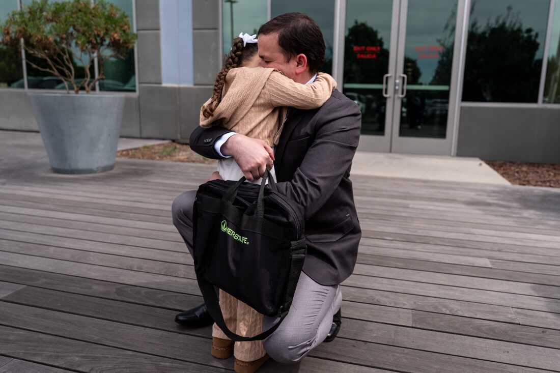Milenko Faria, who is wearing a business suit and carrying a briefcase, is on one knee to hug his young daughter. Her face is not visible.