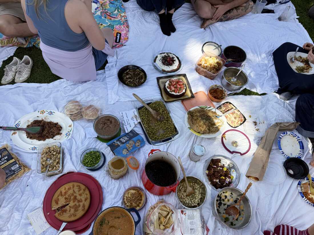 Multiple containers and pots of bean-containing dishes are on white sheets that are spread on the ground. A few people are sitting on the sheets or on the grass near the sheets.