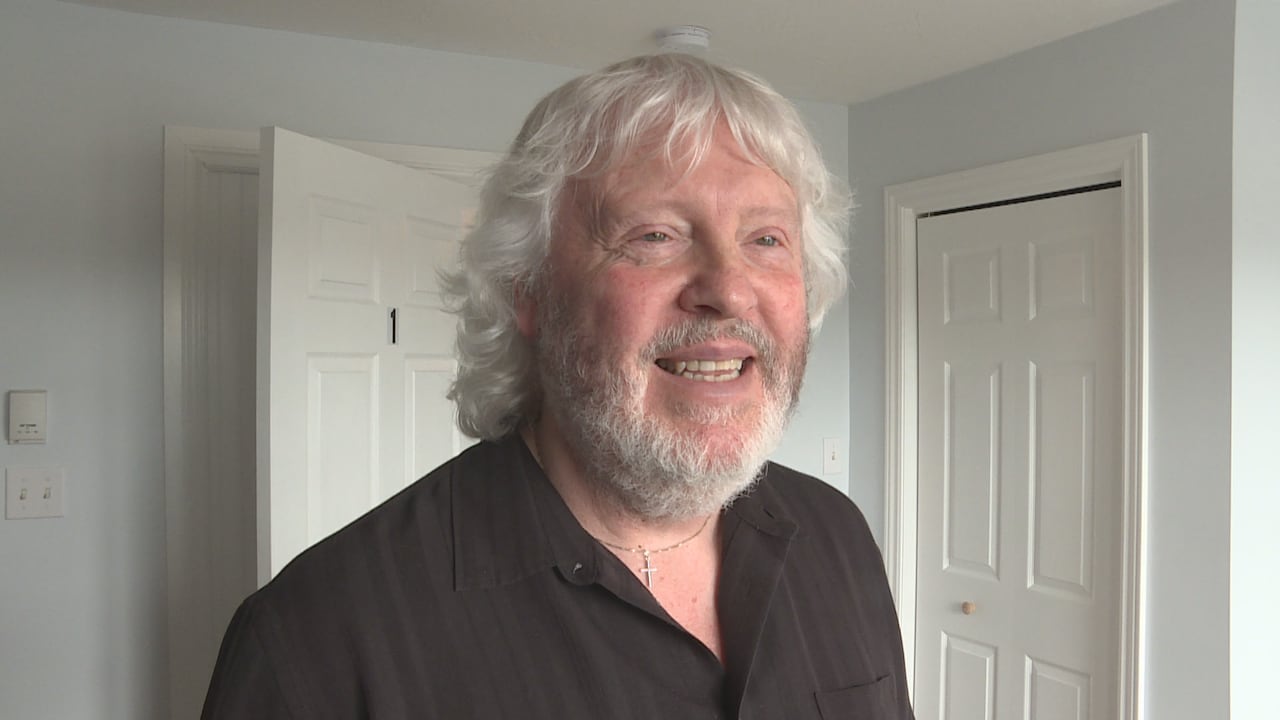 A man with grey hair and a grey beard, wearing a black button up shirt, smiles in a well lit room.