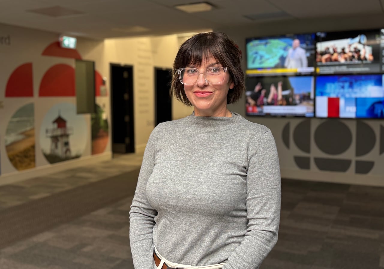 A woman with short, dark hair and clear-framed glasses stands smiling in an office lobby featuring CBC logos and media screens in the background.
