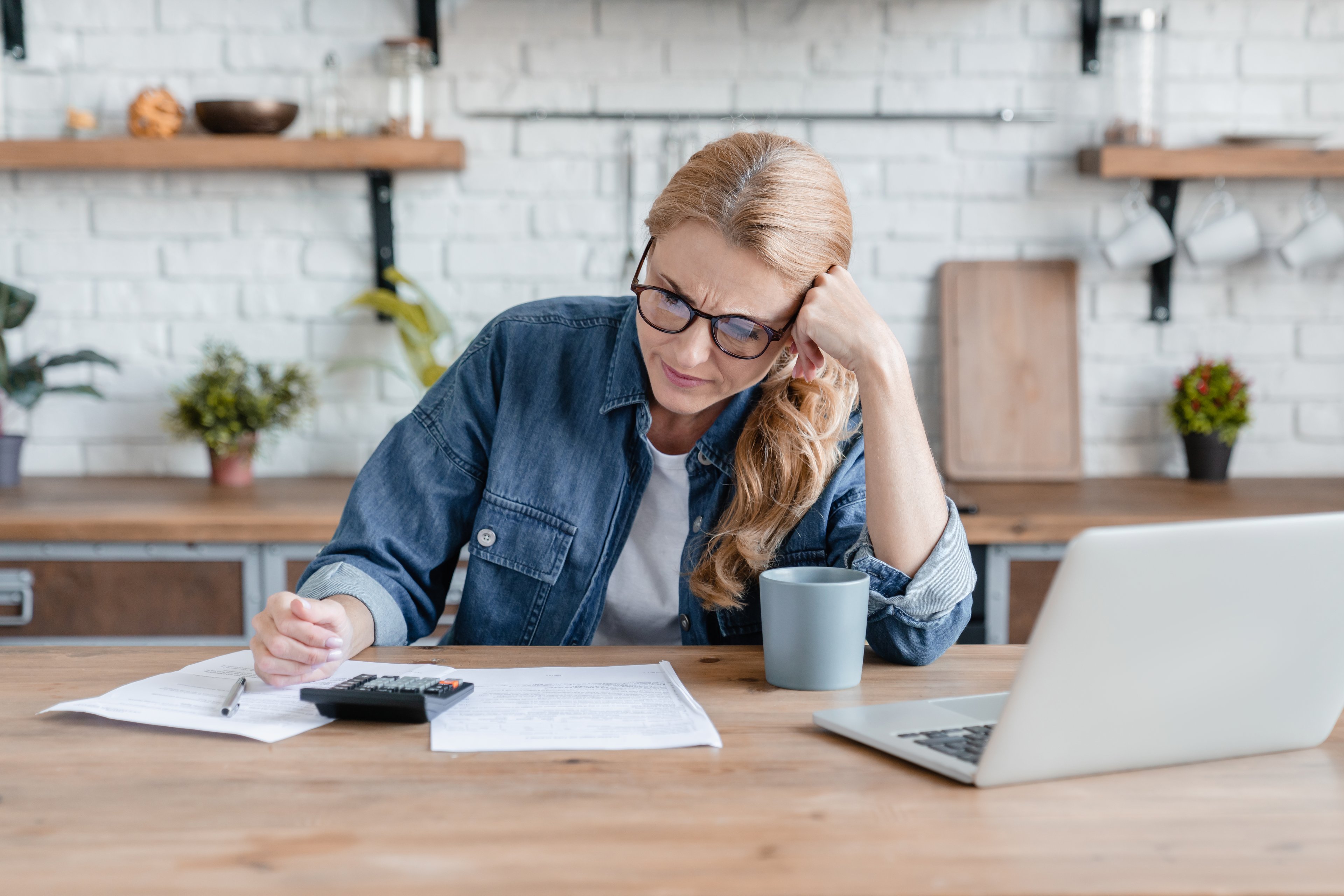 Person sitting at a kitchen table in front of a laptop, reviewing papers.