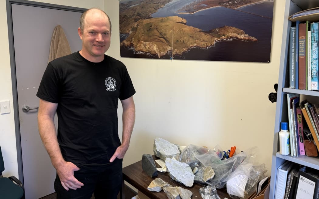 Mid shot of Paul Viskovic looking at camera, he is standing to the left of a table of rock samples on a table.
