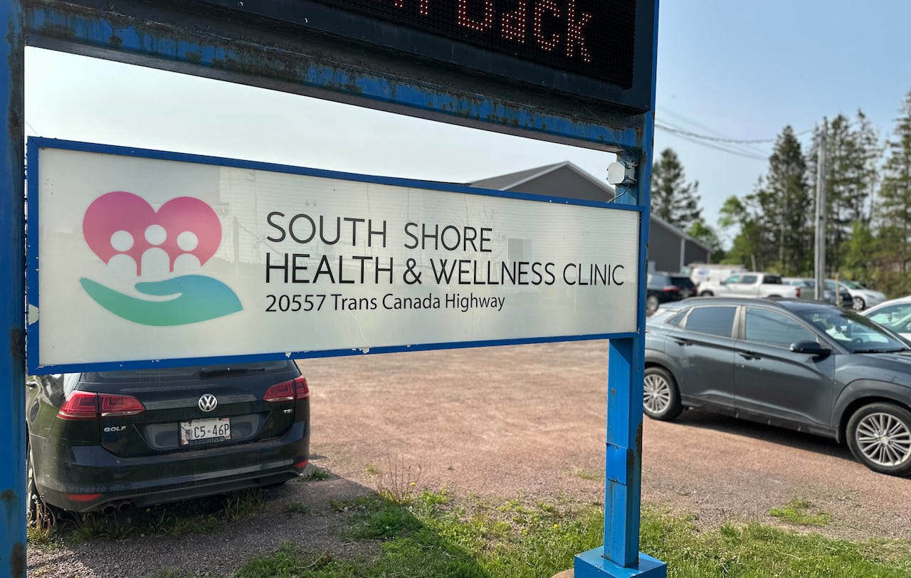 An outdoor close-up shot of a white and blue sign for the "South Shore Health & Wellness Clinic" located at 20557 Trans Canada Highway, with a parking lot and cars visible in the background.