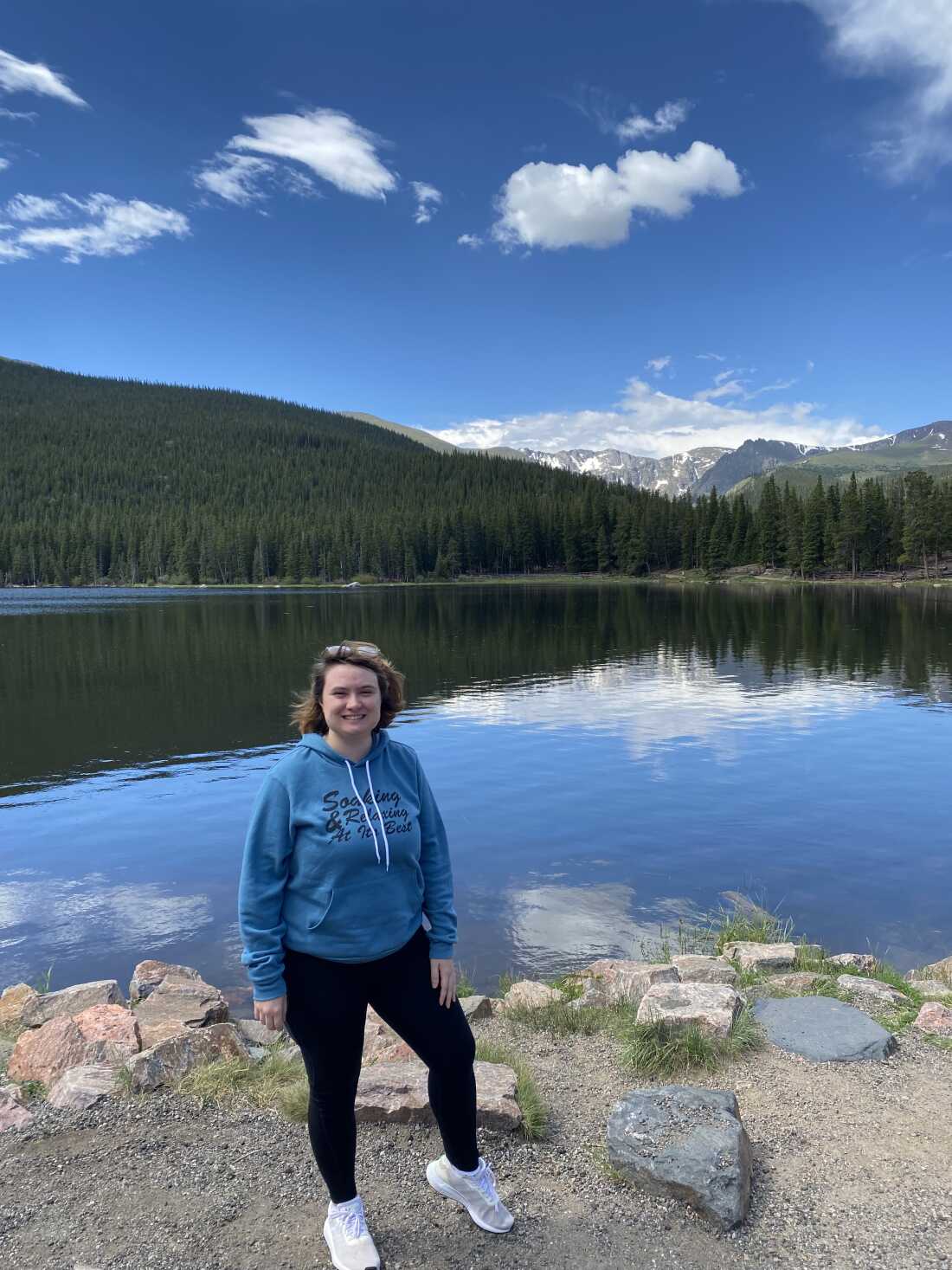 Molly Quinn stands at the shore of a beautiful lake. Pine trees and mountains are in the background.