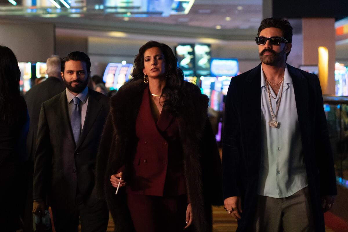 Two men in blazers flank a woman in a maroon suit and fur coat as they walk through a casino floor.