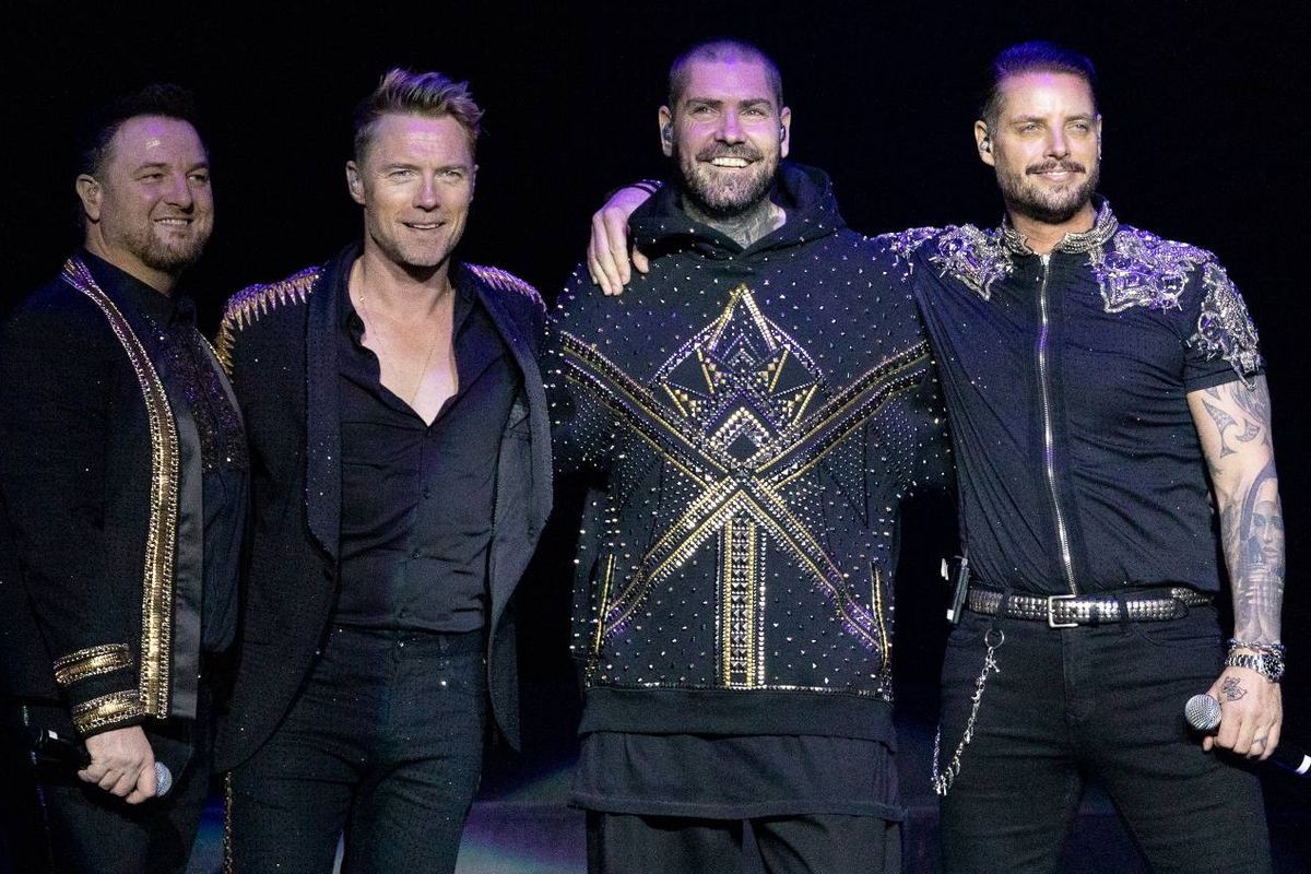 Four individuals dressed in black attire, likely musicians, are standing together in a dimly lit area, possibly on a stage, with one individual prominently positioned in the center.