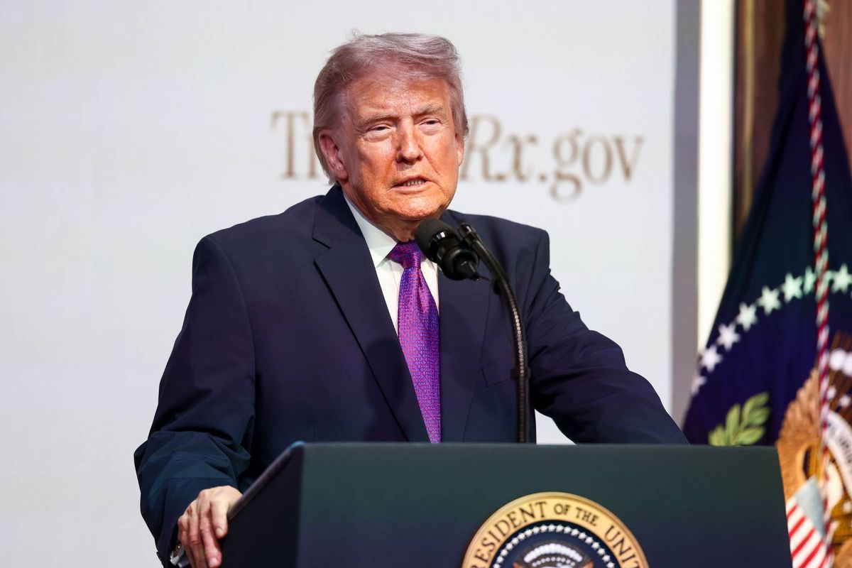 An individual in formal attire, standing at a podium equipped with a microphone, addresses an audience in a formal setting. The backdrop features a white wall adorned with a logo and flag.
