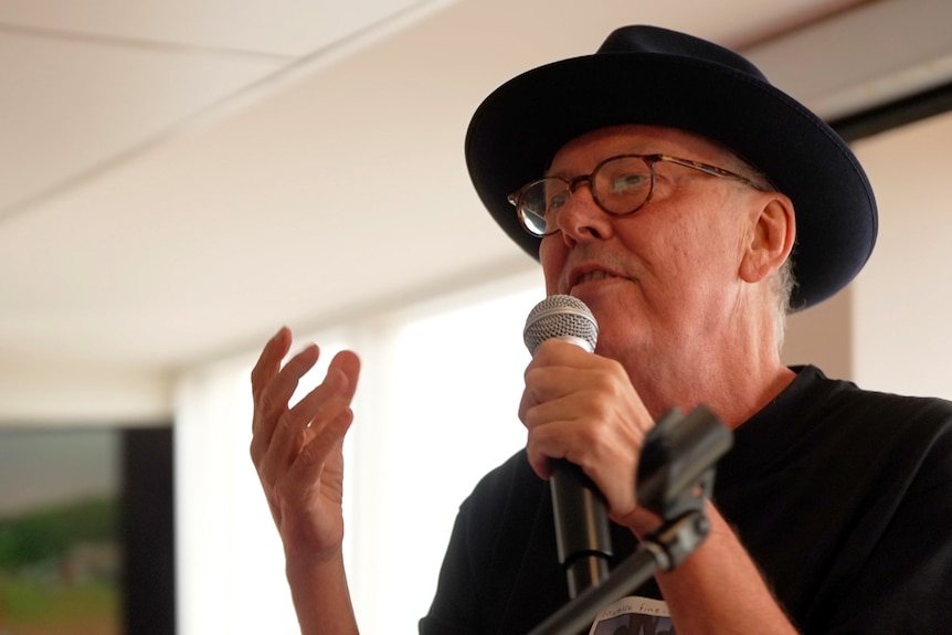 A man wearing a black hat and shirt speaks into a microphone.