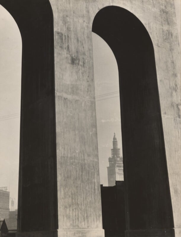 A tall city skyscraper is seen through the large, shadowed arches of a massive concrete structure, creating a dramatic architectural contrast with light and shadow.