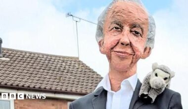 A sculpture depicting Sir David Attenborough wearing a black suit and a white shirt with an animal toy perched on his shoulder. He is perched on a fence which is out of shot. In the background there are some houses.