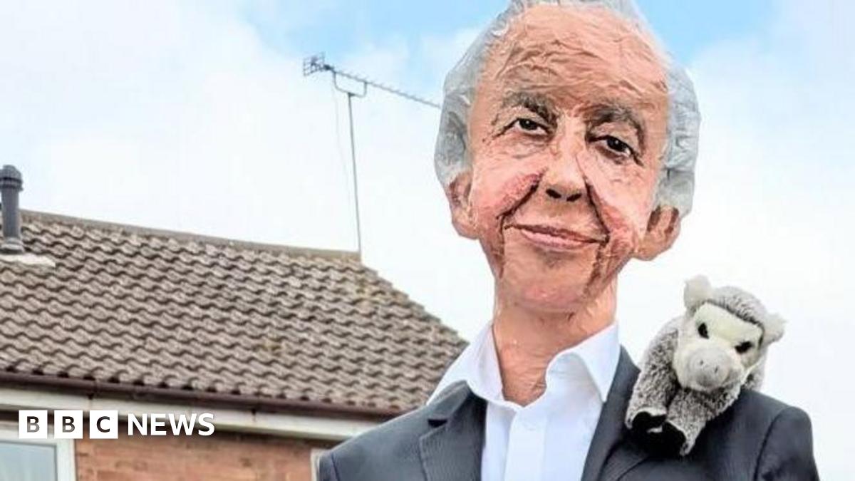A sculpture depicting Sir David Attenborough wearing a black suit and a white shirt with an animal toy perched on his shoulder. He is perched on a fence which is out of shot. In the background there are some houses.
