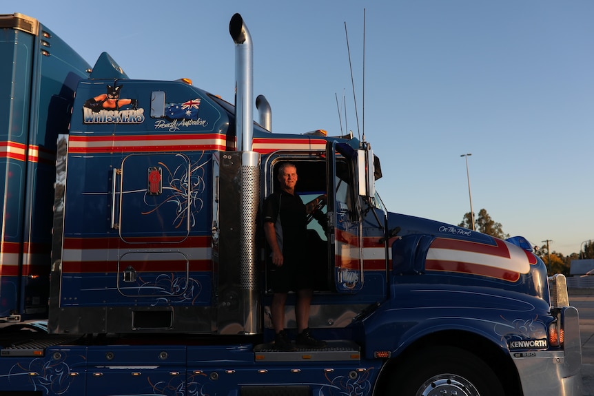 A man stands in the doorway of the cab of his truck as the afternoon sun casts an orange glow.