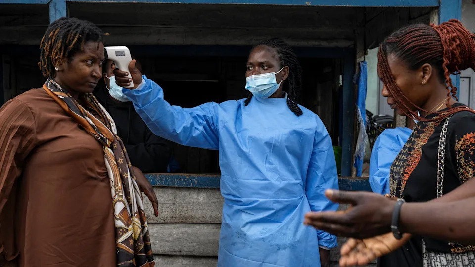 On a street in the DR Congo city of Goma, a health worker in a blue face mask and medical apron takes the temperature of a woman with a device near her head. Another woman and man stand on the other side waiting.