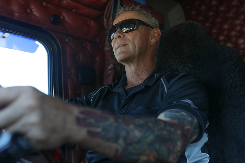 Sean Mulvaney drives a truck with sunglasses on and tattoos seen covering his left arm.