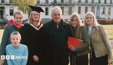 An old photo of a family standing together. Michael Gwilliam is in the middle of his four daughters, with one of them wearing a graduation cap and gown. A young boy crouches down in front of them.