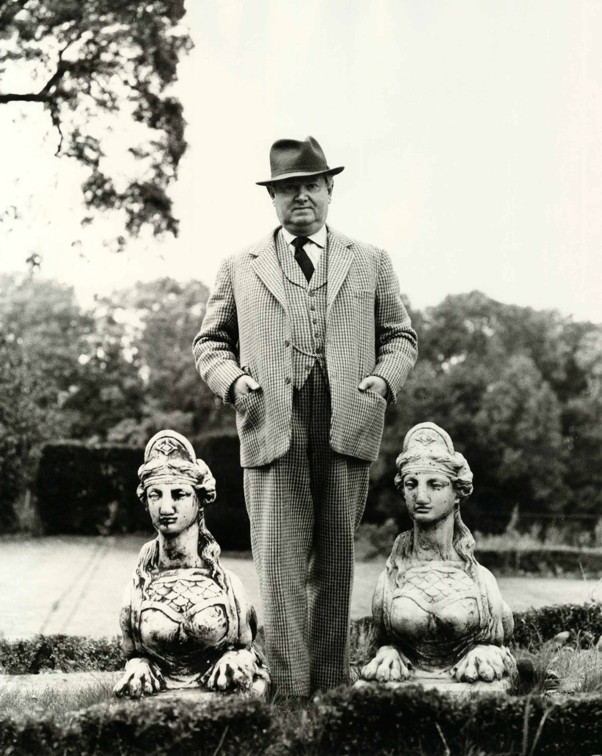 Evelyn Waugh standing between two sphinx statues in a garden.