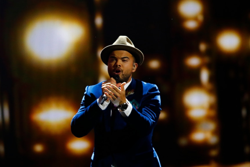 Guy Sebastian singing into a microphone, wearing a blue suit jacket and a fedora-like hat