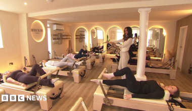A Reformer Pilates studio with seven machines, each one being used by a woman, and a female instructor stood in the middle of the room