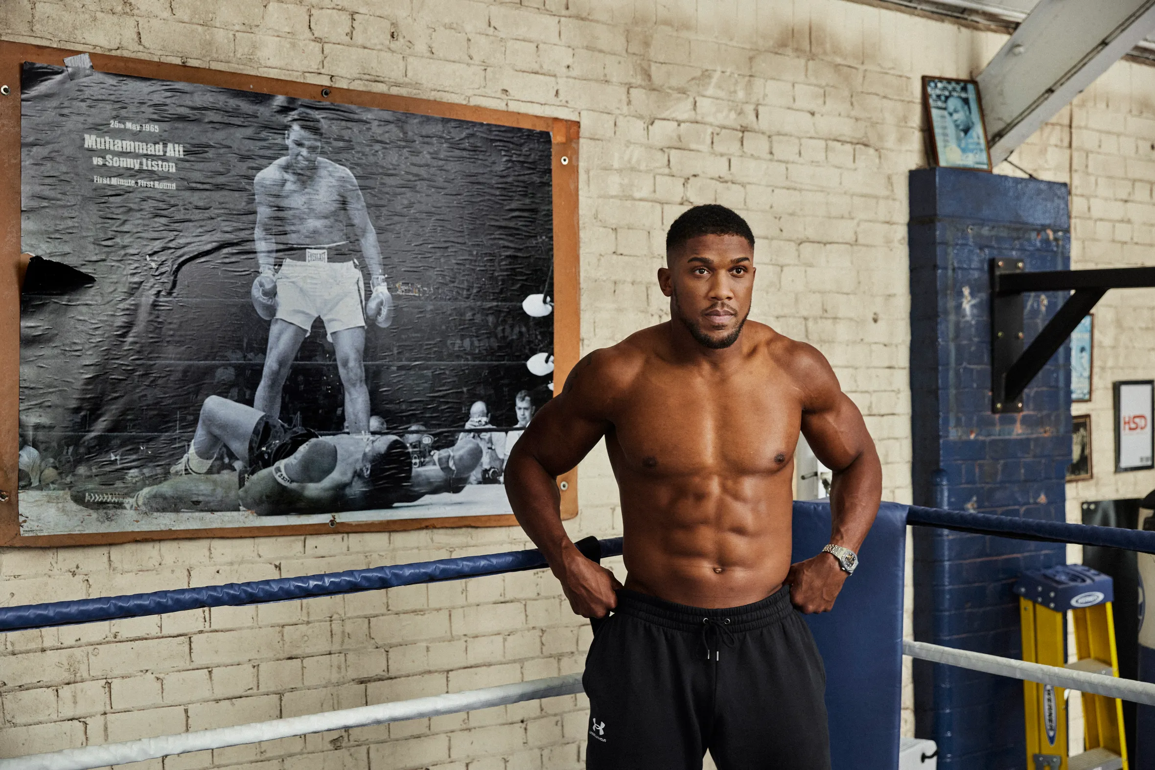 Anthony Joshua, a British heavyweight boxer, shirtless in a boxing ring, with a poster of Muhammad Ali vs Sonny Liston in the background.