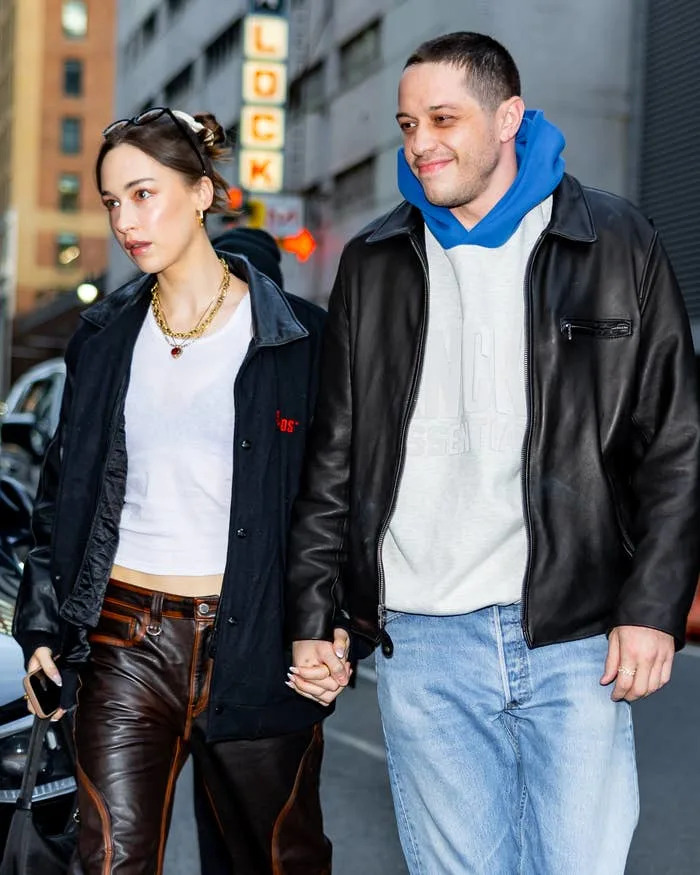 Two people are holding hands on the street. One wears a casual leather jacket and blue jeans, the other stylish layered casual wear