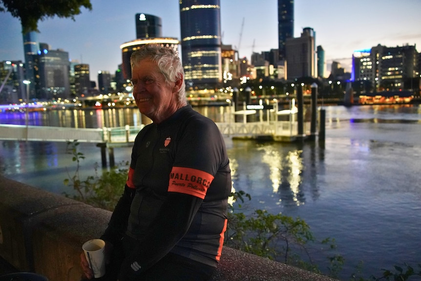 A man leans against a wall next to the Brisbane River with the city skyline behind him.