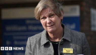 A blonde woman in a grey jacket and a yellow name tag with the name "Tracey" written on it.