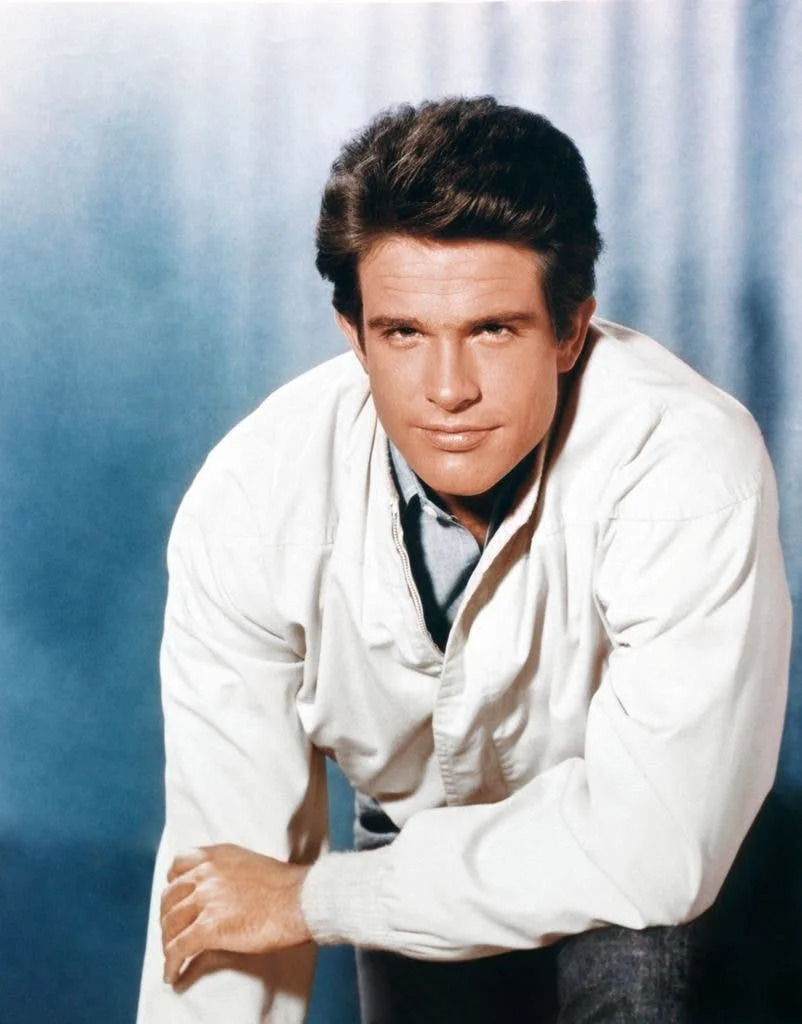 Warren Beatty in a white jacket, shirt and jeans, resting on one arm and holding his forearm with his other hand.