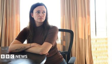 Claire Brosseau is photographed at her home in Toronto, Canada. She has dark brown hair and blue eyes. She is seated at a table with her arms crossed. She is wearing a brown short-sleeved turtle neck.