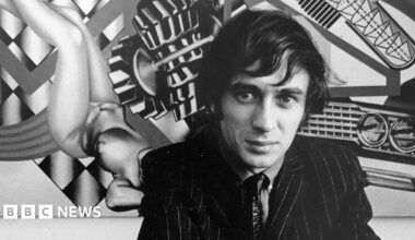 A black and white image of Peter Phillips in front of one of his distinctive artworks. He is wearing a dark pinstriped suit, tie and shirt. He is crouching down in front of the painting.