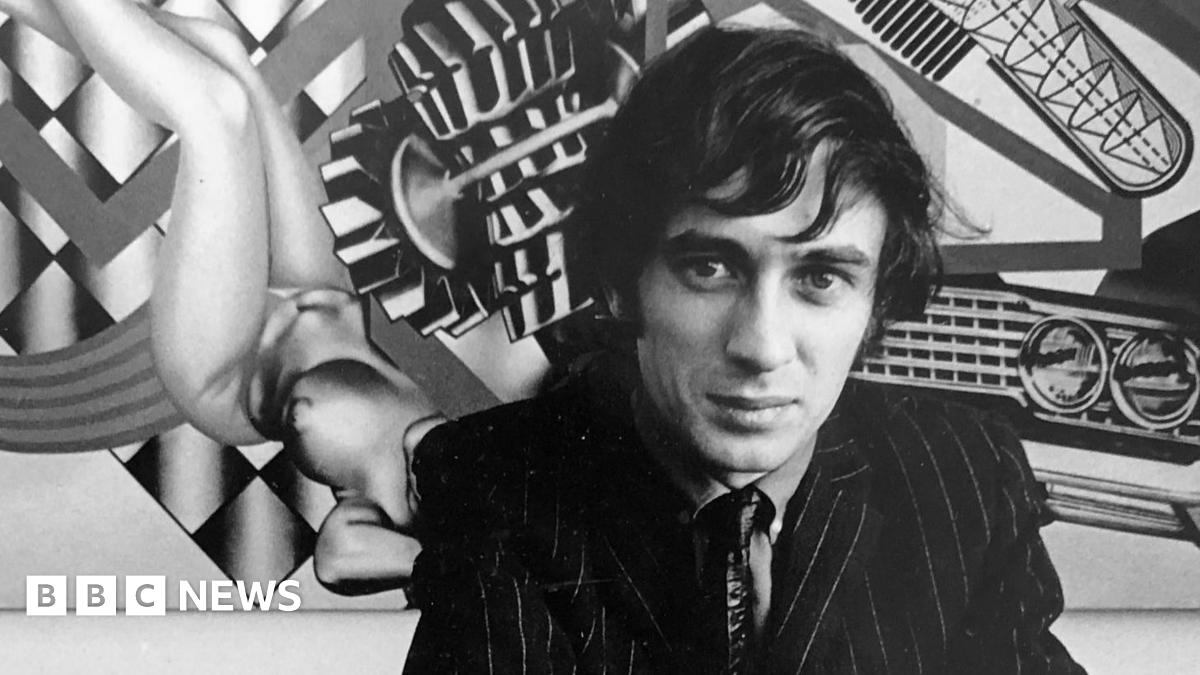 A black and white image of Peter Phillips in front of one of his distinctive artworks. He is wearing a dark pinstriped suit, tie and shirt. He is crouching down in front of the painting.