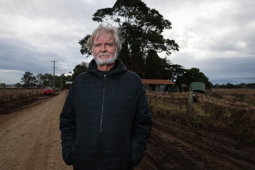 A middle aged man wearing a fleece jacket stands on a gravel road outside a country home, on a cloudy day, looking serious.