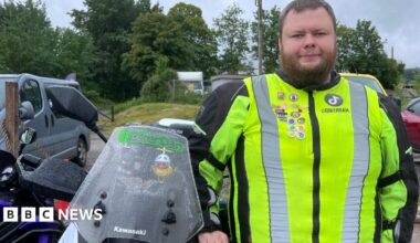A man with a beard in a high vis jacket stands next to a motorbike in a car park with trees in the background. He has "BikerDan" written on the windshield of his motorbike.
