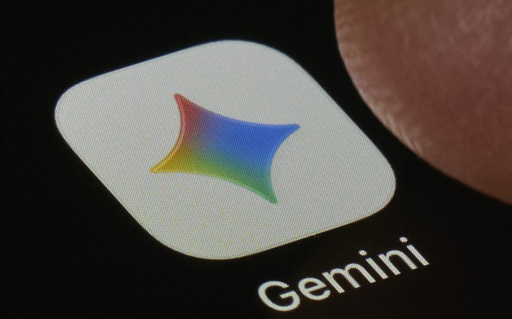 A macro close-up photograph shows the Google Gemini AI application icon on a smartphone screen set against a dark background in San Ferdinando di Puglia, Italy, on May 17, 2026. The icon displays a colorful four-pointed star with a gradient ranging from red and orange through blue and green, positioned on a white rounded-rectangle background. The Gemini wordmark appears in white below the icon. A fingertip is partially visible at the upper right. Gemini is Google's flagship generative AI assistant, representing a major competitor in the consumer-facing large language model market as of May 2026. (Photo Illustration by Matteo Della Torre/NurPhoto) (Photo by Matteo Della Torre / NurPhoto via AFP)