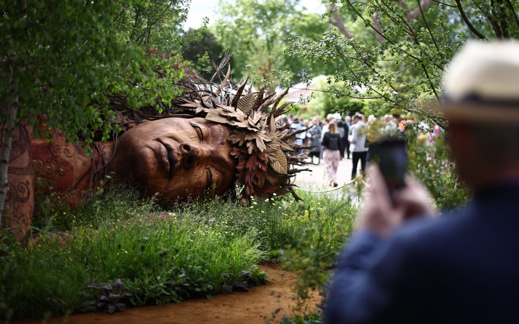 A visitor takes a photo of the Gaia in The Campaign to Protect Rural England's Garden: "On the Edge" on the press day ahead of the 2026 RHS Chelsea Flower Show in London on May 18, 2026. Emphasising nature's power to protect, a giant fallen tree in one garden has been carved into a sculpture of a sleeping guardian, Gaia, or Mother Nature. "The garden aims to bring to you the potential and the joy in the ordinary," said Sarah Eberle, designer of the Garden on the Edge from the Campaign for Protection of Rural England. (Photo by Henry Nicholls / AFP)