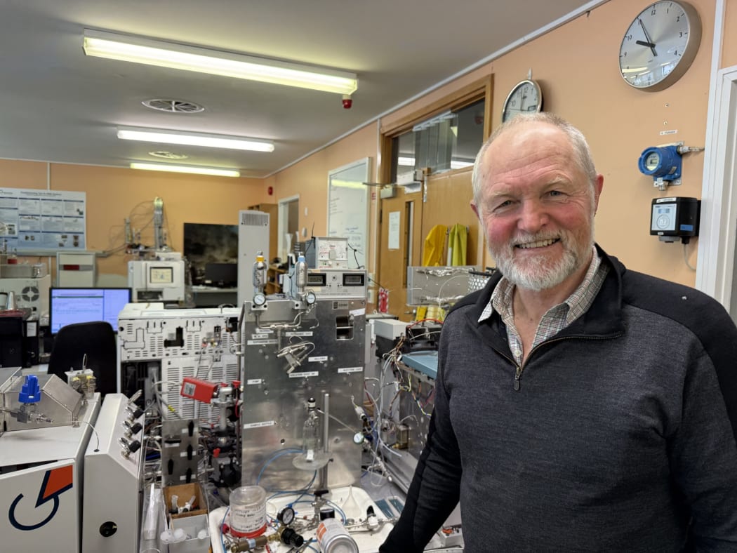 Mid shot of Kevin Faure smiling at camera, standing in National Isotope Centre lab, with stainless equipment and wires in the background