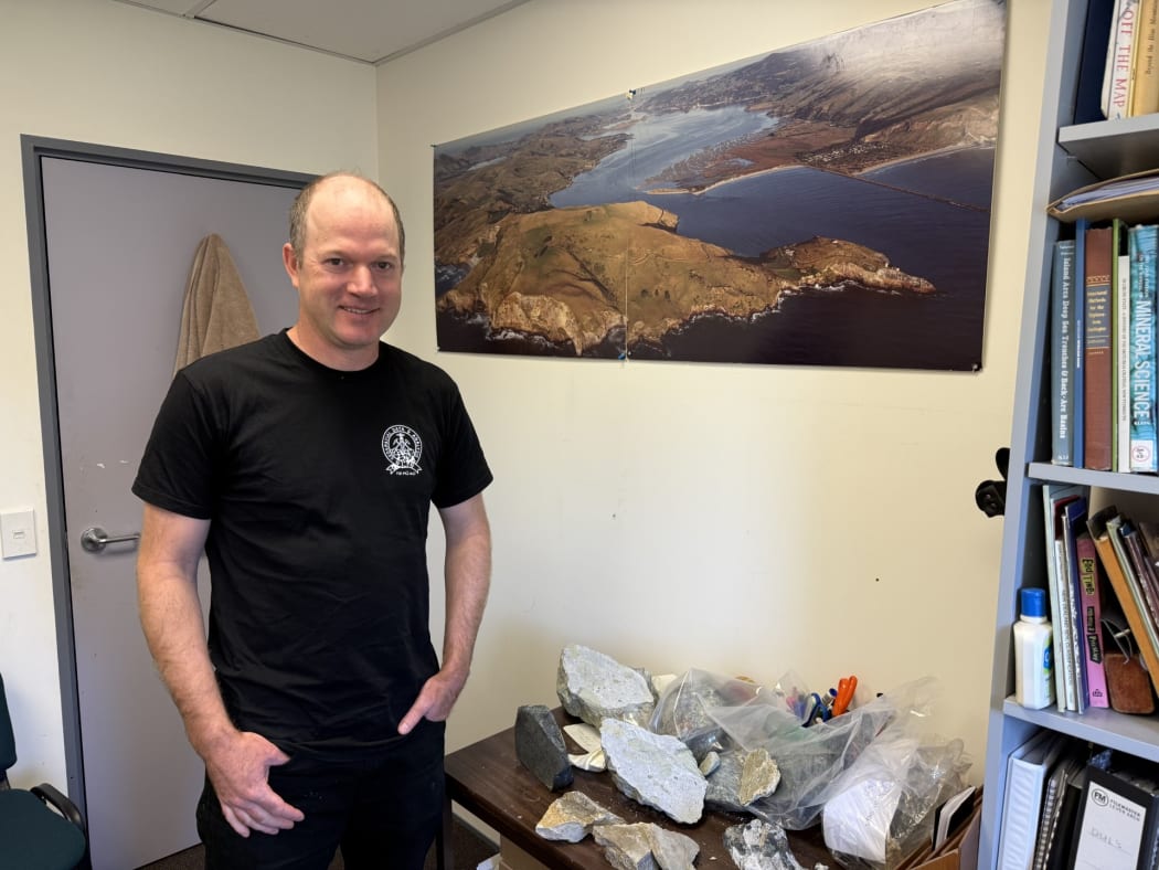Mid shot of Paul Viskovic looking at camera, he is standing to the left of a table of rock samples on a table.