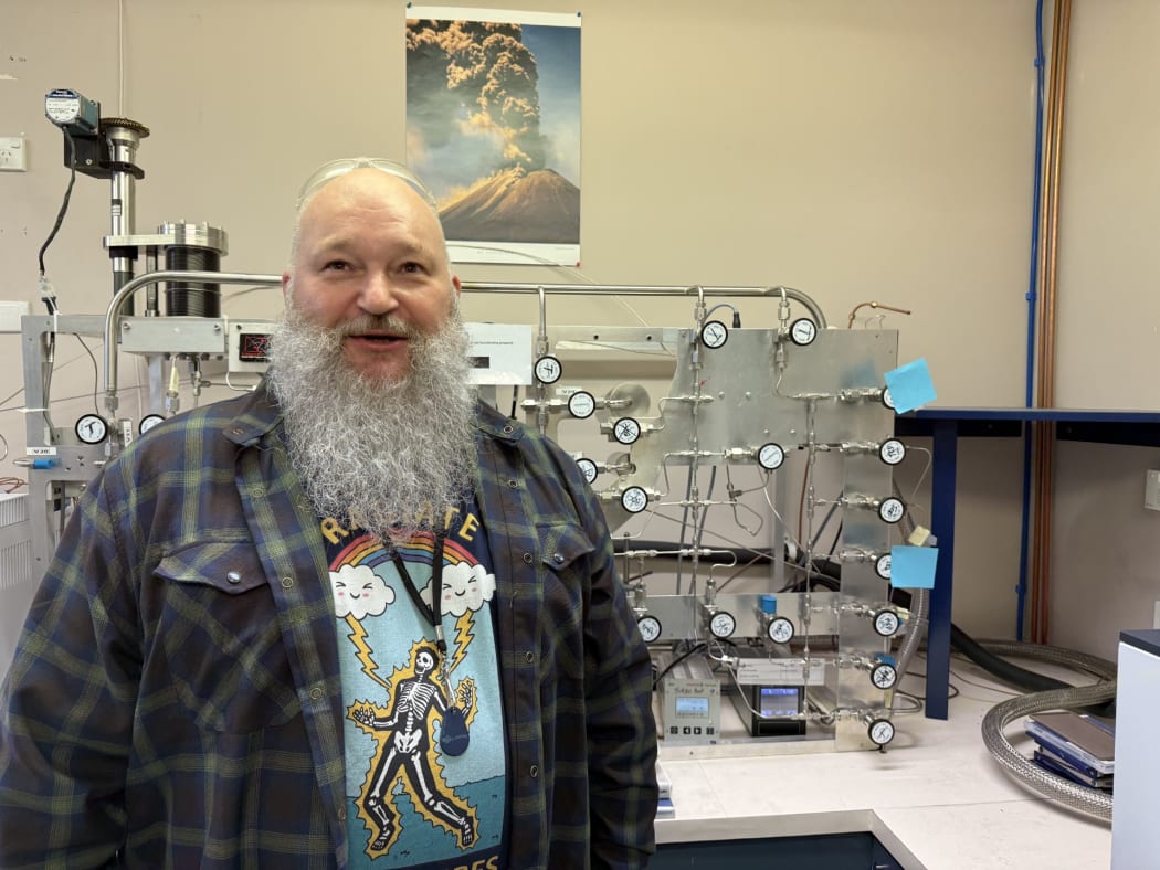 Mid shot of isotope hydrogeologist Thijs van Soest, standing in a lab looking at the camera. Behind him to the right a vacuum manifold sits on a bench, a complex of silver metal pipes and gagues