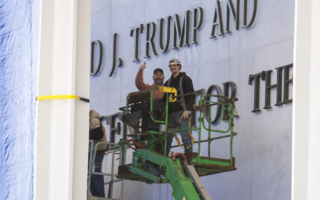 Workers react to the media as they come down off a scissor lift after updating signage outside the The John F. Kennedy Memorial Center for the Performing Arts after the building was renamed in honor of US President Donald Trump on December 19, 2025, in Washington, DC. The US capital's renowned John F. Kennedy arts center was renamed the "Trump-Kennedy Center" after President Donald Trump, the White House announced on December 18, 2025. Press Secretary Karoline Leavitt said the center's board -- which Trump purged of Democrats earlier this year -- voted for the change because of the Republican's "unbelievable work" in overhauling the famed venue. (Photo by Jim WATSON / AFP)