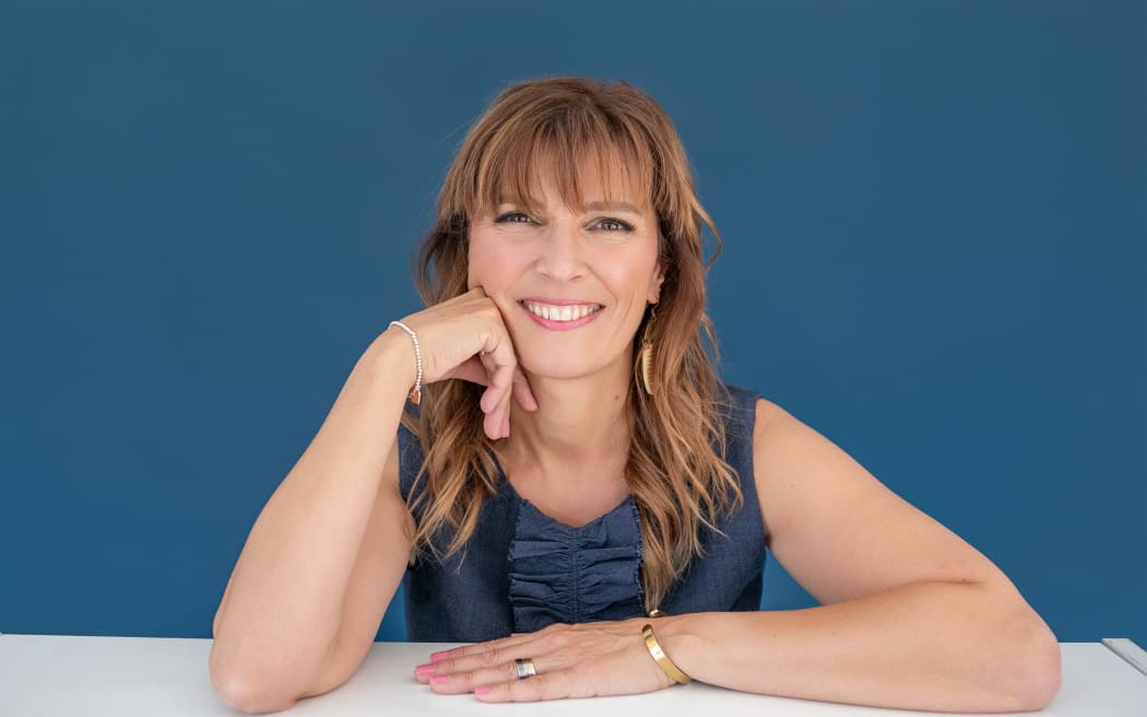 Sarah Davis is sitting behind a white desk and smiling at the camera. She is wearing a blue top that is a slightly darker shade than the blue wall behind her.