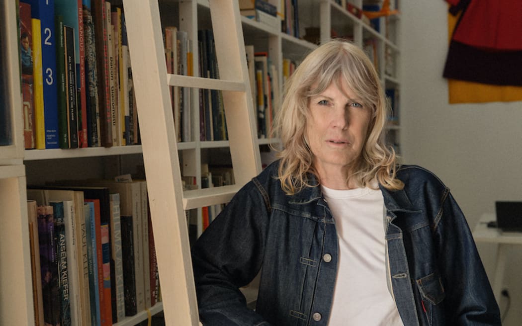 Judy Millar is in what looks like a library. There is a wall of books to the left. She is leaning on one of those ladders that helps people get to high shelves. There is what appears to be a painting of a red dress on the wall behind her.