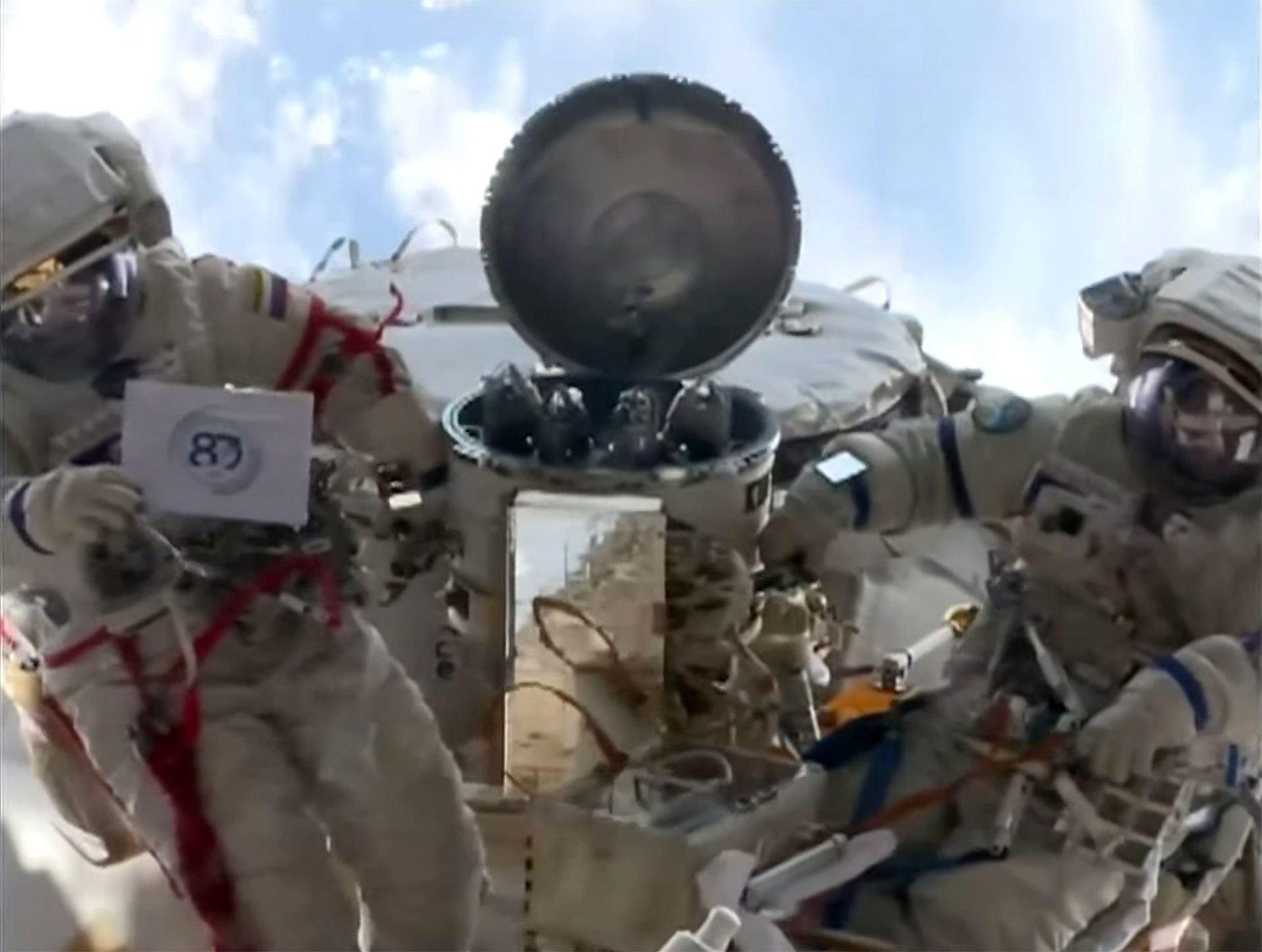 Two cosmonauts pose for photographs with a small sign during a spacewalk outside of a space station in Earth orbit.