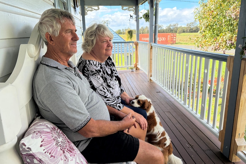 Couple in their 70s with their little brown and white lap dog looking up at them sitting looking out from their front verandah.