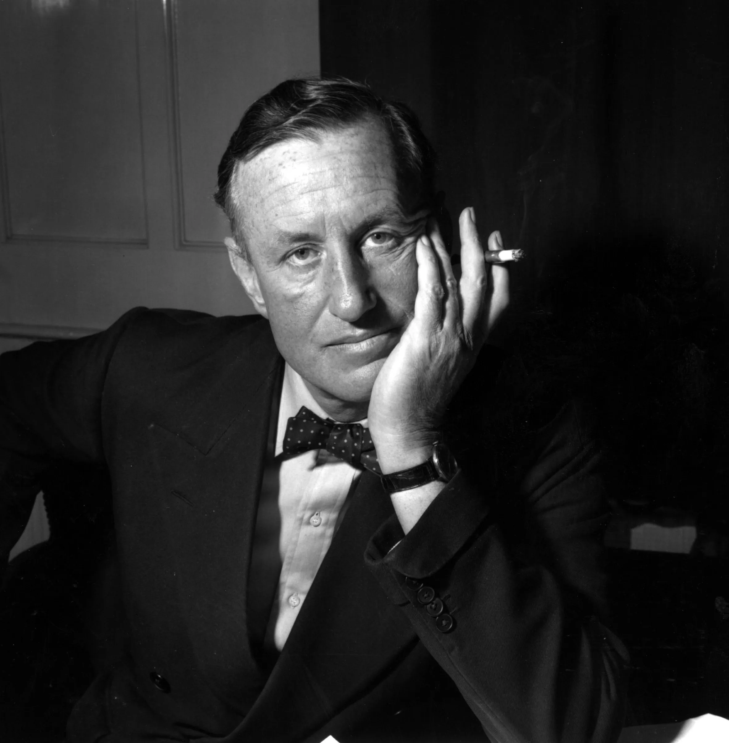Ian Fleming, creator of James Bond, sits with his hand on his face and a lit cigarette between his fingers.