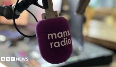 A microphone in a radio studio which has a purple pop shield over it with Manx Radio in white lettering across its face.