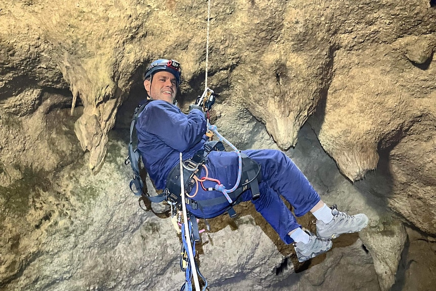 A man in blue coveralls, a helmet with a light and wearing a harness is dropped into a void with a cave roof background.