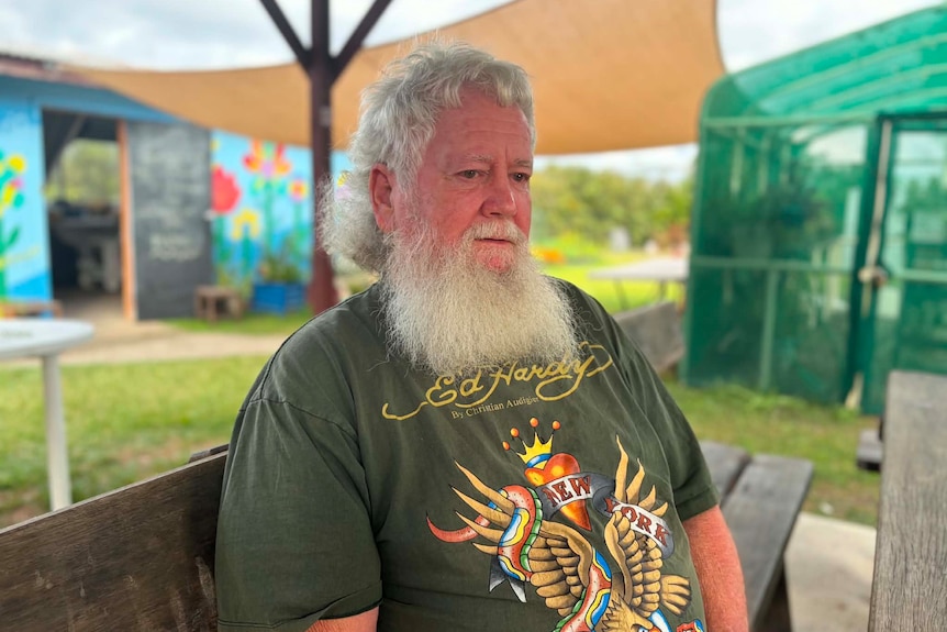A man with white hair and beard sits at an outdoor table looking down with shed and a greenhouse behind him
