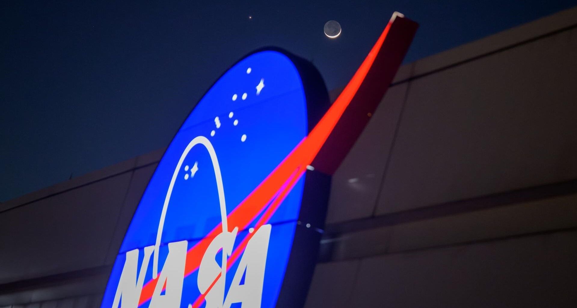 The crescent moon and Venus shine in the night sky above the NASA "Meatball" logo on the side of a building.