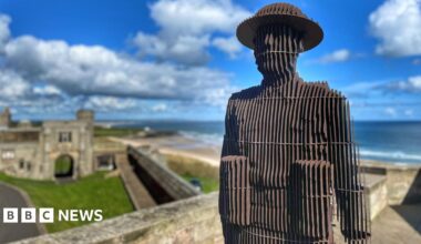 Sculpture of soldier constructed from vertical steel plates standing on a castle wall with the sea in the background.