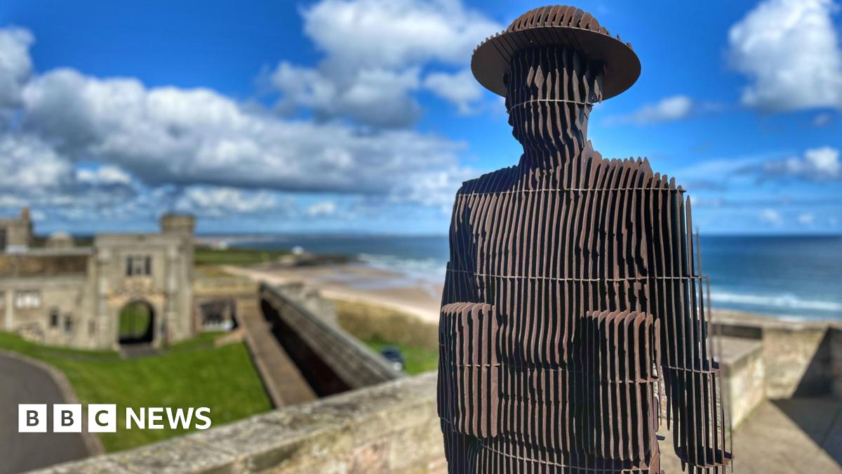 Sculpture of soldier constructed from vertical steel plates standing on a castle wall with the sea in the background.