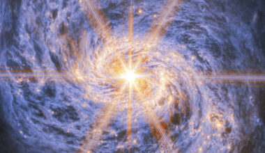 A spiral galaxy shown in mid-infrared light. The image is dominated by an extremely bright glow from the galaxy’s nucleus. Six large and two smaller rays of light emit from the center, which are diffraction spikes created by the telescope’s optics. The galaxy’s spiral arms are visible by two lines of glowing orange bubbles which whirl out into the disc. Swirling blue clouds of dust make up the rest of the galaxy.